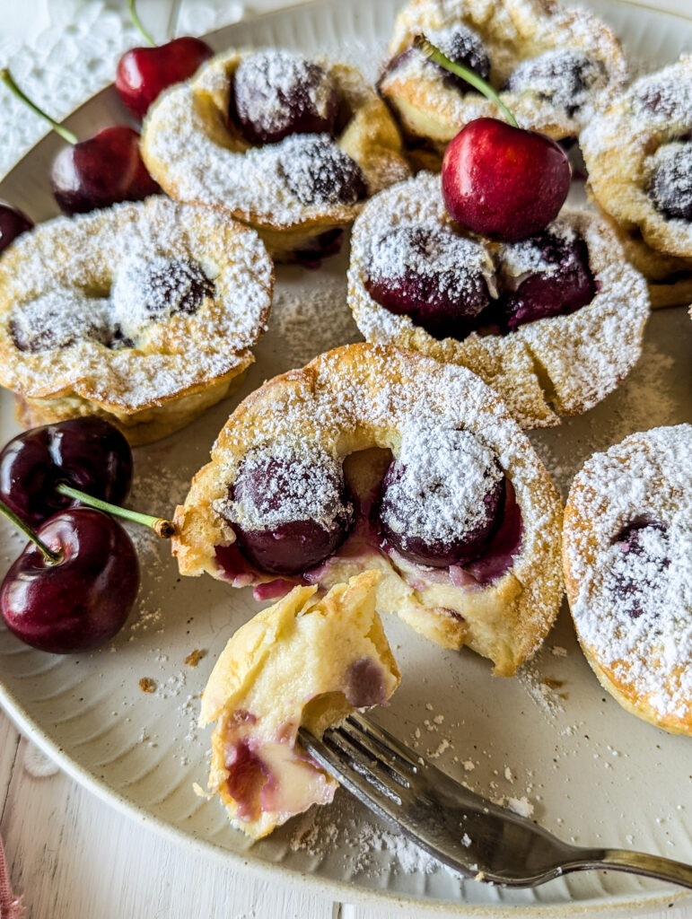 Eine Gabel mit einem Stück vom Mini Clafoutis mit Kirschen. Im Hintergrund einige Mini Clafoutis und ein paar frische Kirschen