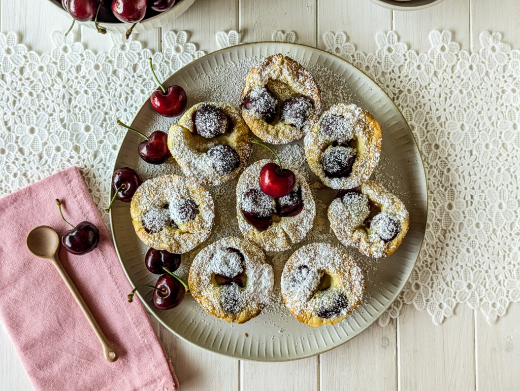 Ein Teller mit Mini Clafoutis - französischen Törtchen aus der Muffinform - garniert mit frischen Kirschen und Puderzucker