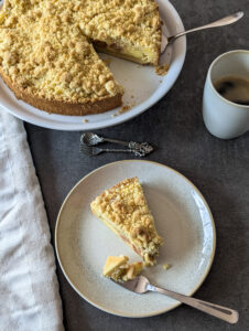 Rhabarber Streuselkuchen mit cremiger Puddingfüllung – ein Stück angeschnitten auf einem Teller, mit sichtbaren Schichten aus Mürbteig, Pudding und knusprigen Streuseln