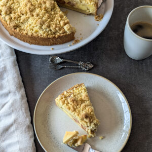 Rhabarber Streuselkuchen mit cremiger Puddingfüllung – ein Stück angeschnitten auf einem Teller, mit sichtbaren Schichten aus Mürbteig, Pudding und knusprigen Streuseln