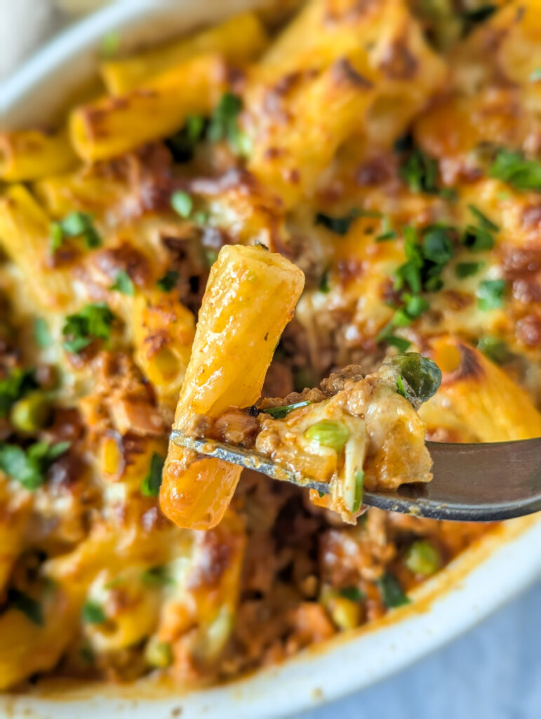 Nudel auf einer Gabel mit Käse, Hackfleisch und einer cremigen Soße. Im Hintergrund Rigatoni al forno in der Auflaufform