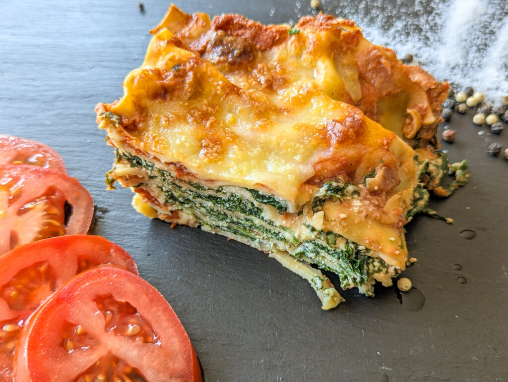 Saftige Spinat-Ricotta-Lasagne auf einem schwarzem Teller mit frischen Tomaten und Pfefferkörnern garniert. Die Käsekruste ist goldbraun und die Spinat-Nudel-Schicht ist sichtbar.