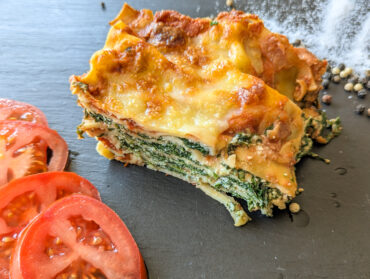Spinat Ricotta Lasagne auf einem schwarzem Teller mit Tomaten und Pfefferkörner. Die Kruste ist goldbraun und die Nudel-Spinat-Schichten sind deutlich zu sehen