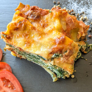 Ein Stück Spinat-Ricotta-Lasagne auf einem schwarzen Teller. Die Lasagne ist goldbraun überbacken und die Nudelschichten und der Spinat sind deutlich sichtbar