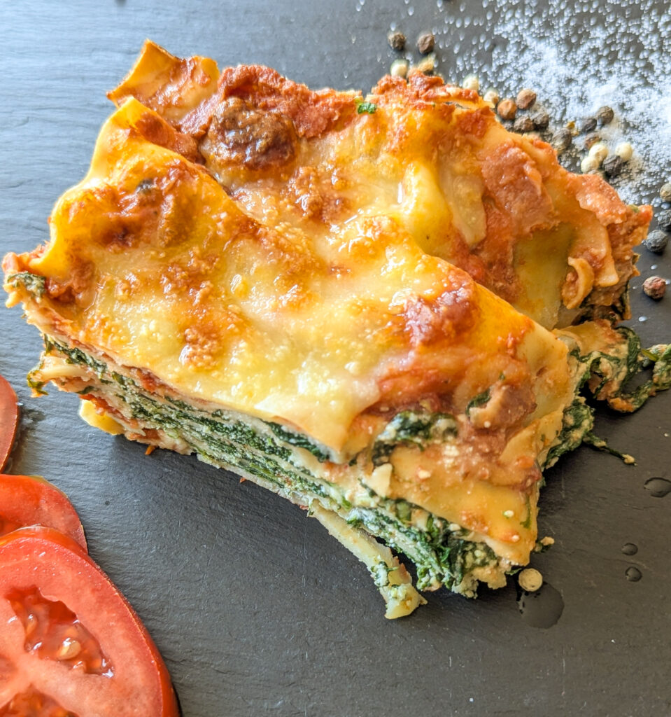Ein Stück Spinat-Ricotta-Lasagne liegt auf einem schwarzen Teller. Die Lasagne ist goldbraun und knusprig und die Spinat-Nudel-Schichten sind deutlich sichtbar.