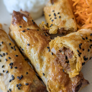 Knusprige Stangen Börek mit Hackfleisch auf einem Teller, bestreut mit Schwarzkümmel, daneben Tzatziki und Karottensalat