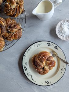 Eine gedeckte Kaffeetafel mit einem Teller voller Kanelbullar aufeinandergestapelt - davor liegt ein Kanelbullar auf einem hellen Teller mit einem silbernen Löffel