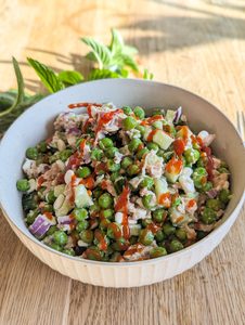 Erbsensalat mit Hüttenkäse, Tunfisch und Gurken und roten Spritzern Sriracha Soße in einer hellen Schüssel